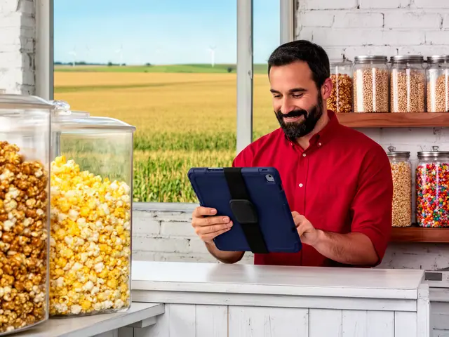 Bearded man in a red shirt checking a navy tablet behind the counter of a popcorn shop with big glass bins